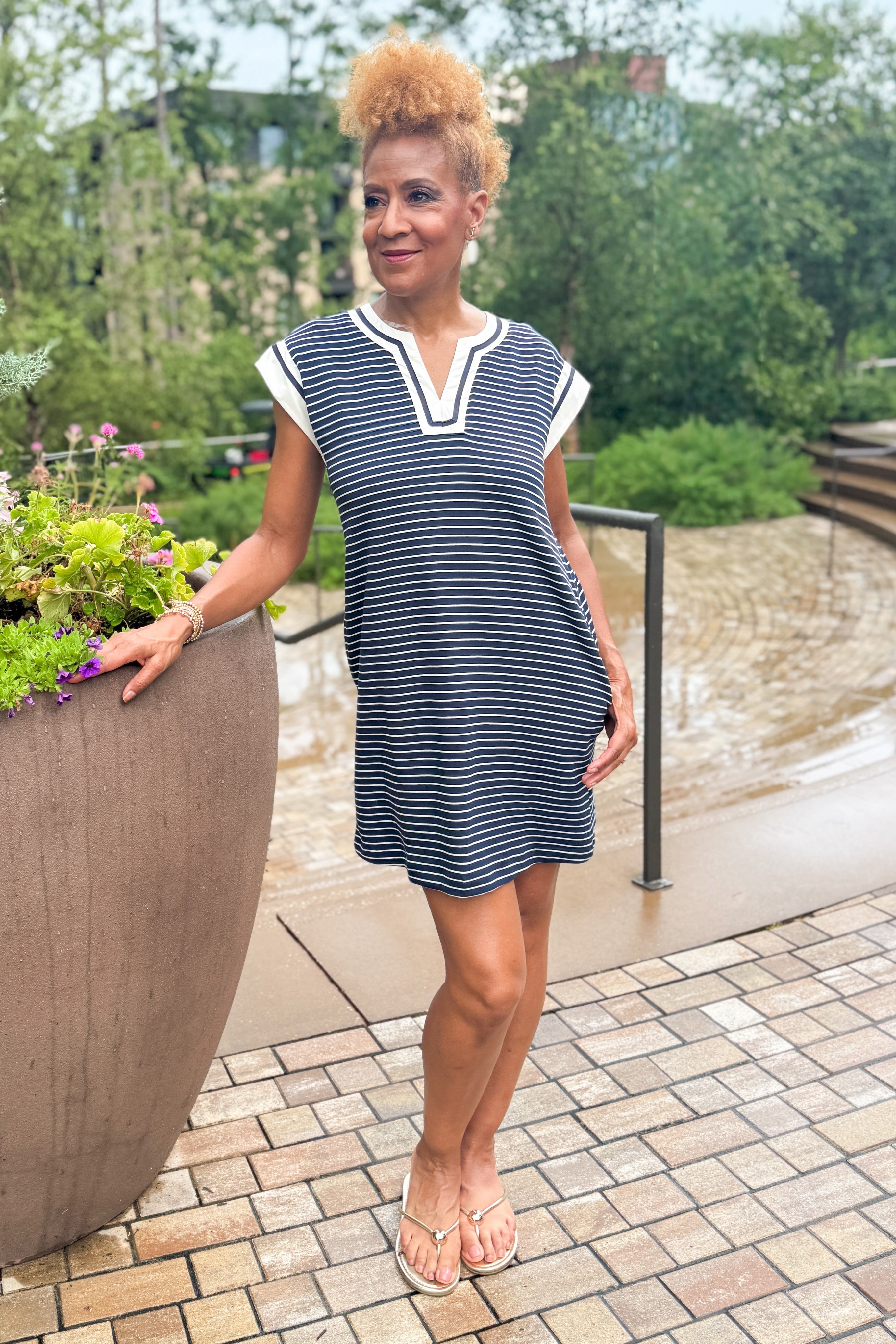 Front view of woman wearing Swagger Boutique’s Navy Striped V-Neck Shift Dress with sandals standing outside by flowers at a park.