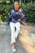 Front view of woman wearing Swagger Boutique’s Navy & Brown Printed Sweater by THML with jeans sitting on a stone wall outside.