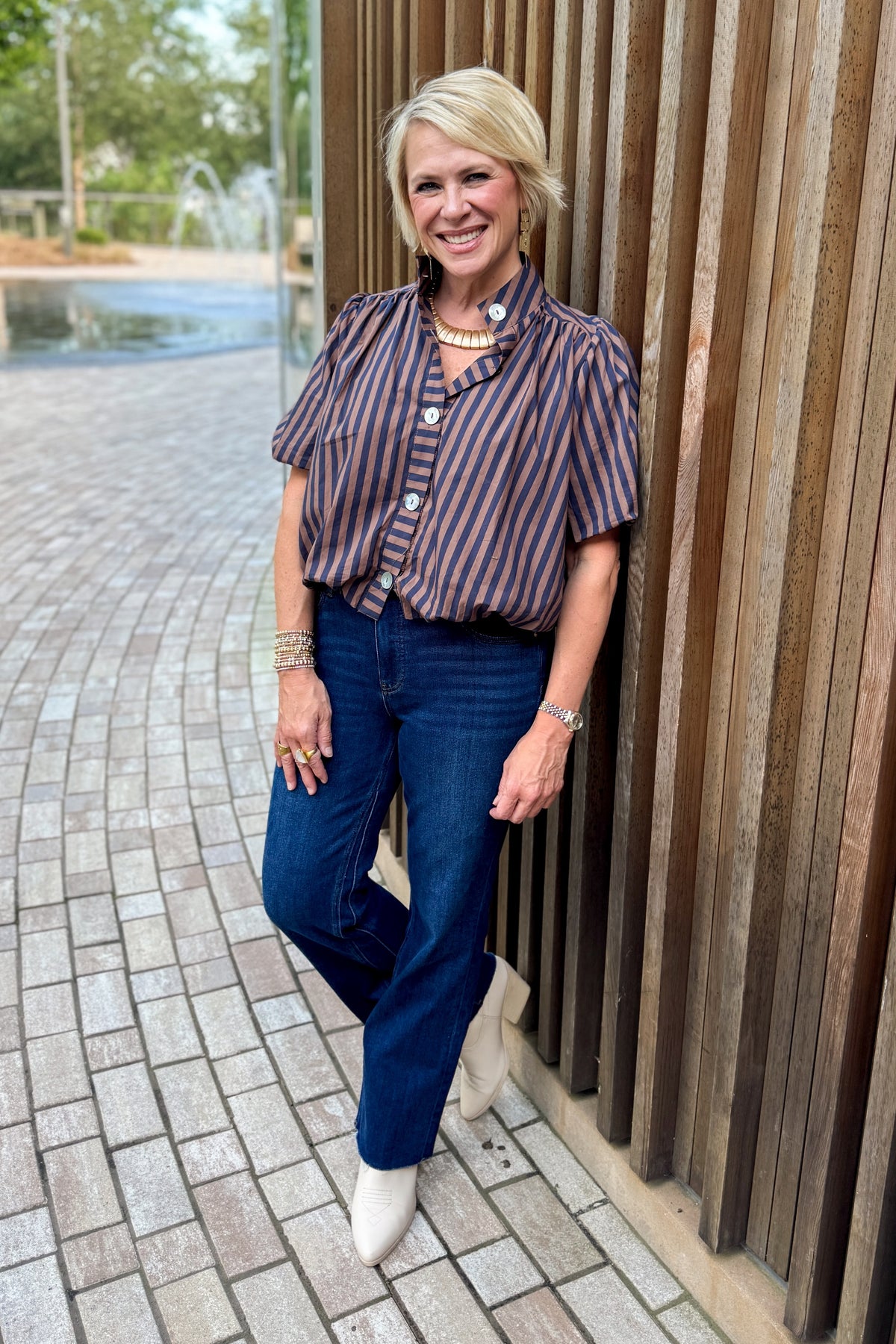 Front view of woman wearing Swagger Boutique’s Navy &amp; Brown Striped Bubble Hem Top and jeans standing outside by a fountain.