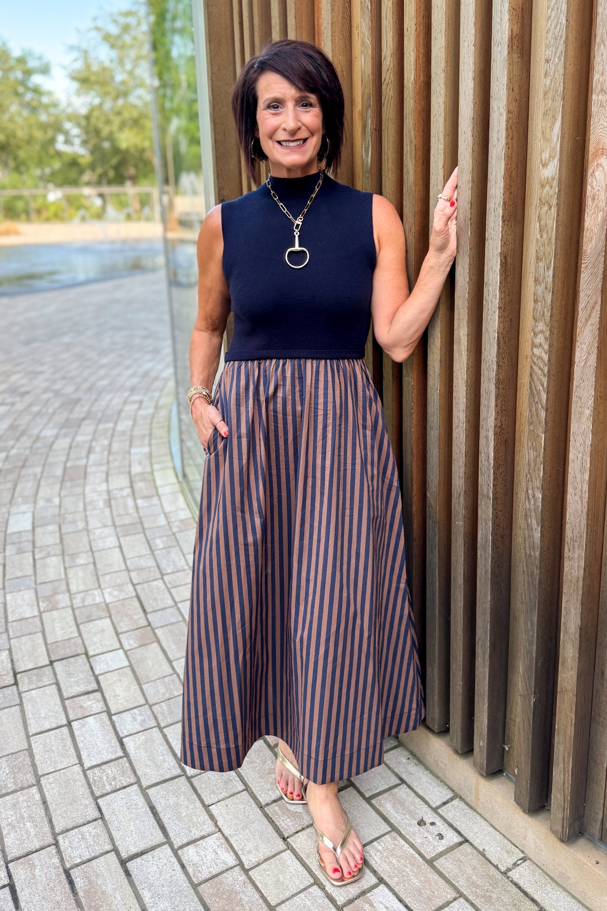Front view of woman wearing Swagger Boutique’s Navy &amp; Brown Striped Layered Mock Neck Midi Dress standing by a fountain outside.