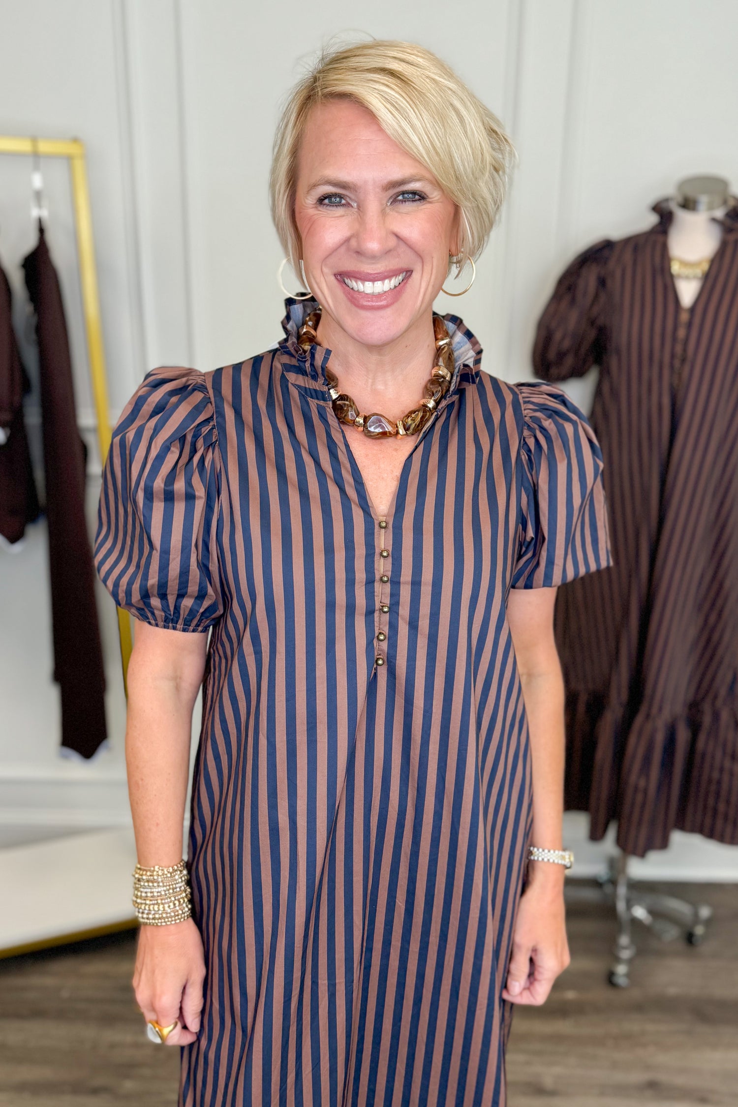 Front view of woman wearing Swagger Boutique’s Navy &amp; Brown Striped Puff Sleeve Midi Dress by Entro with clothing hanging in the background.