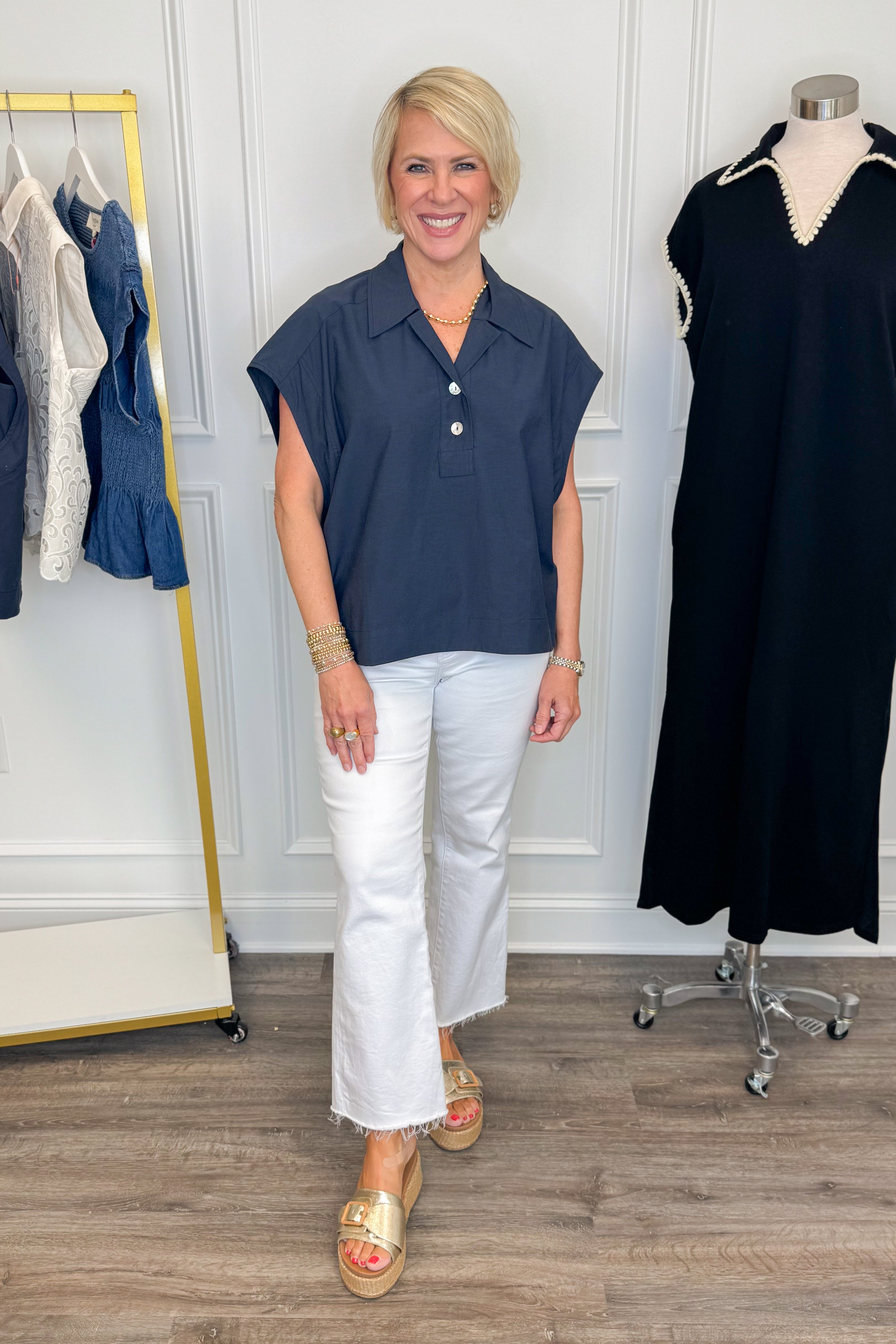 Front view of woman wearing Swagger Boutique’s Navy Collared Cap Sleeve Top  with jeans with clothing hanging in the background.