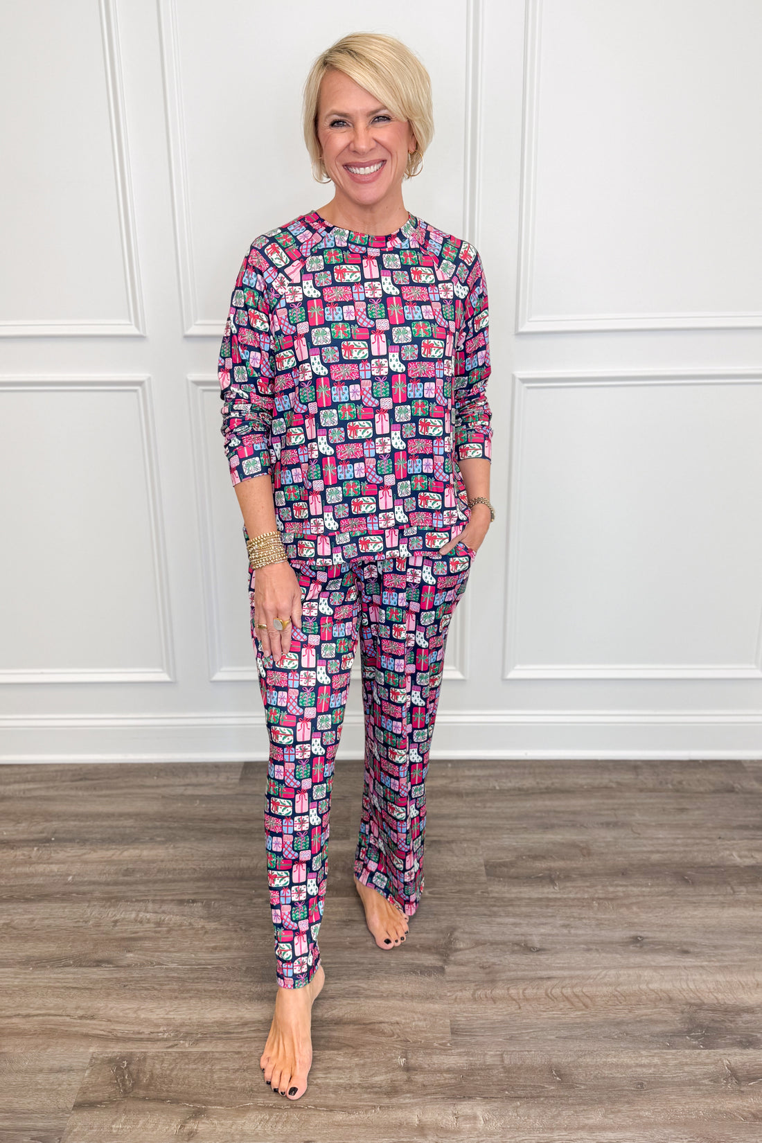 Model wearing the No Peeking Navy Annie Pajama Set, featuring a colorful Christmas present print in pink, green, blue, and white on a navy background. The set includes a relaxed-fit, long-sleeve crewneck top and matching straight-leg pajama pants with side pockets.