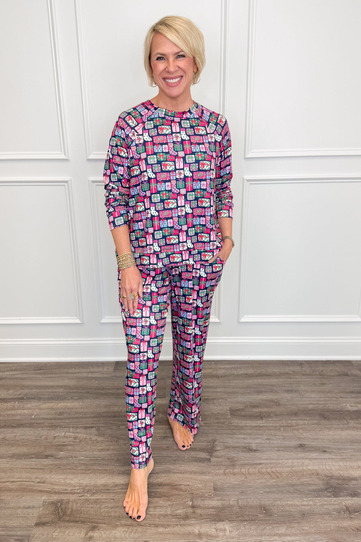 Model wearing the No Peeking Navy Annie Pajama Set, featuring a colorful Christmas present print in pink, green, blue, and white on a navy background. The set includes a relaxed-fit, long-sleeve crewneck top and matching straight-leg pajama pants with side pockets.