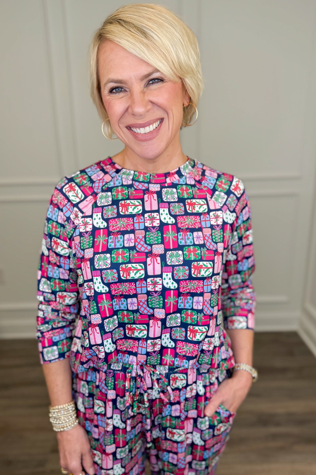 Model wearing the No Peeking Navy Annie Pajama Set, featuring a colorful Christmas present print in pink, green, blue, and white on a navy background. The set includes a relaxed-fit, long-sleeve crewneck top and matching straight-leg pajama pants with side pockets.