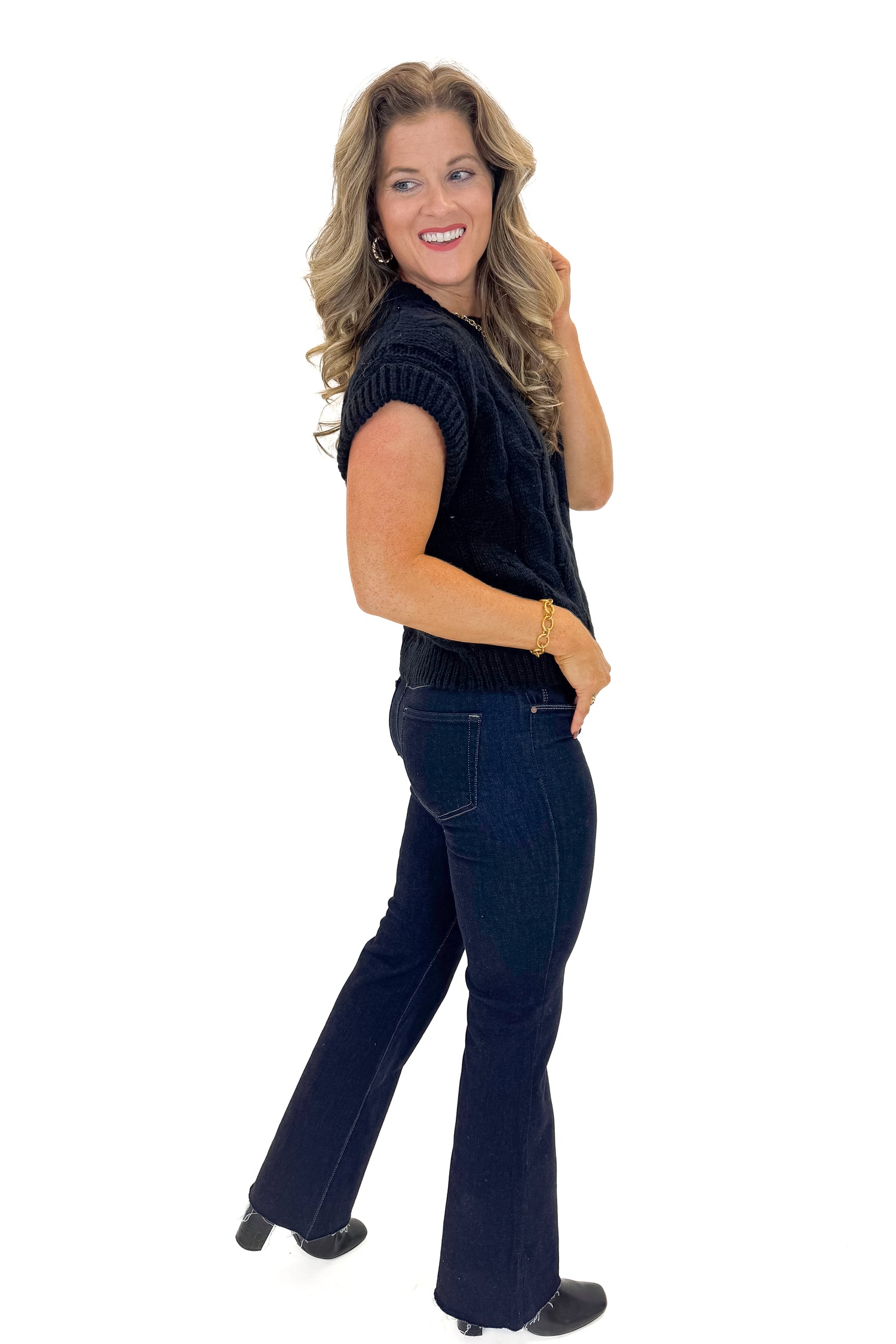 Side view of woman wearing Black Cable Knit Short Sleeve Sweater with jeans against white studio background.