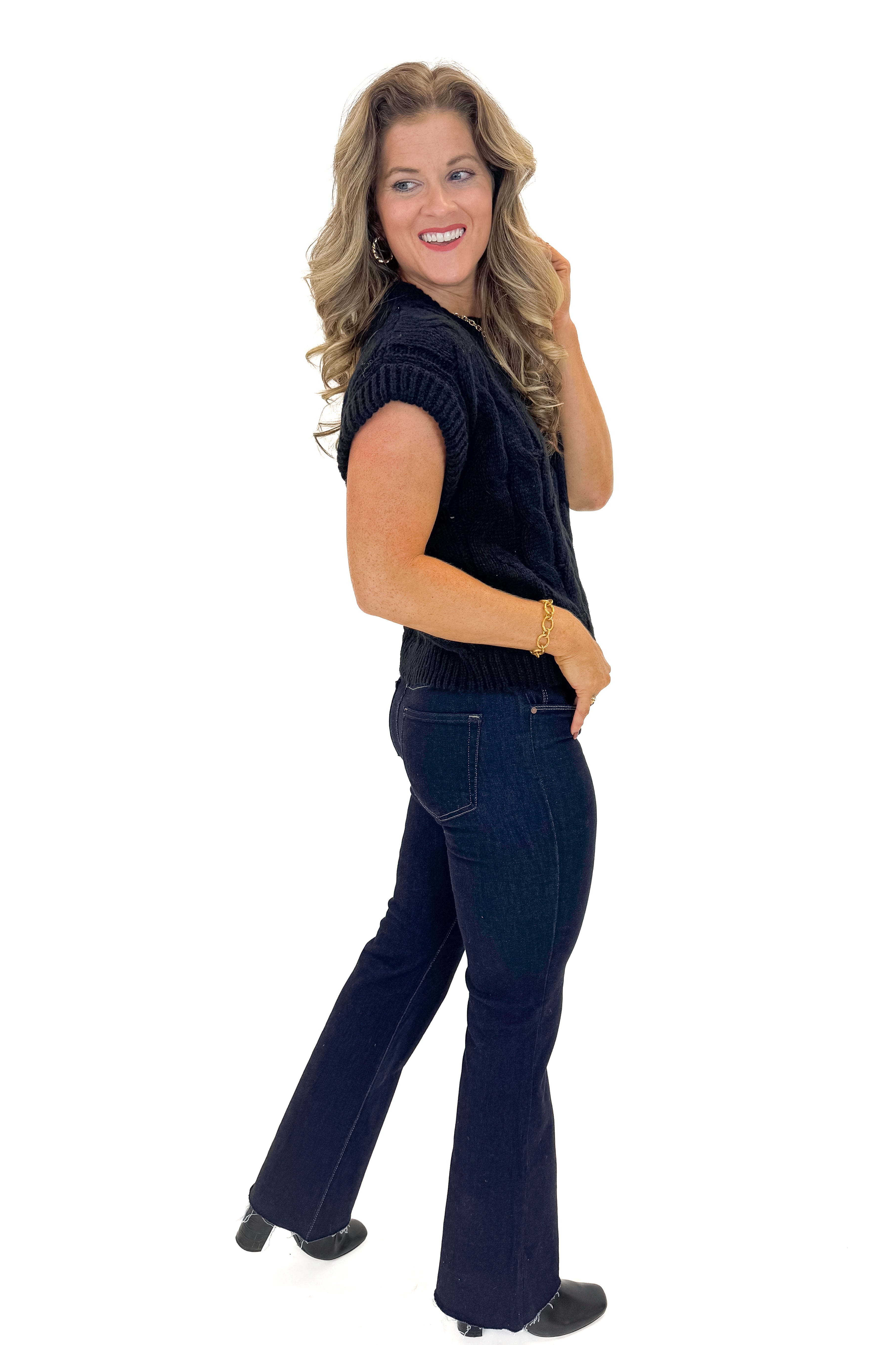 Side view of woman wearing Black Cable Knit Short Sleeve Sweater with jeans against white studio background.