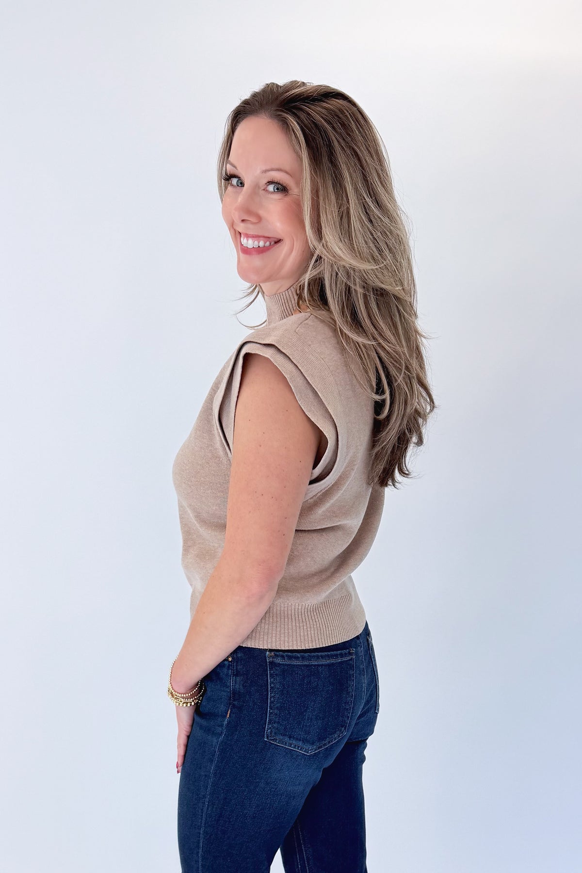 Side view of woman wearing Swagger Boutique’s Oatmeal Mock Neck Sleeveless Sweater by Entro with jeans against white studio background.