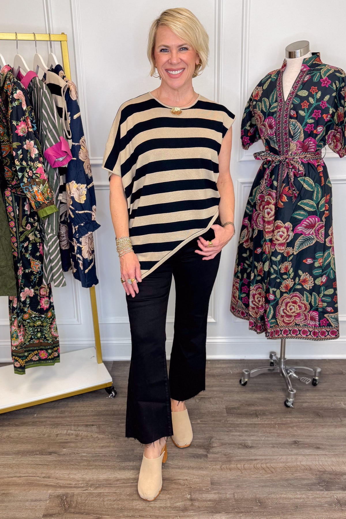 Front view of woman wearing Swagger Boutique’s Oatmeal &amp; Black Striped Asymmetrical Top with jeans with clothing hanging in the background.
