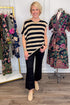 Front view of woman wearing Swagger Boutique’s Oatmeal & Black Striped Asymmetrical Top with jeans with clothing hanging in the background.