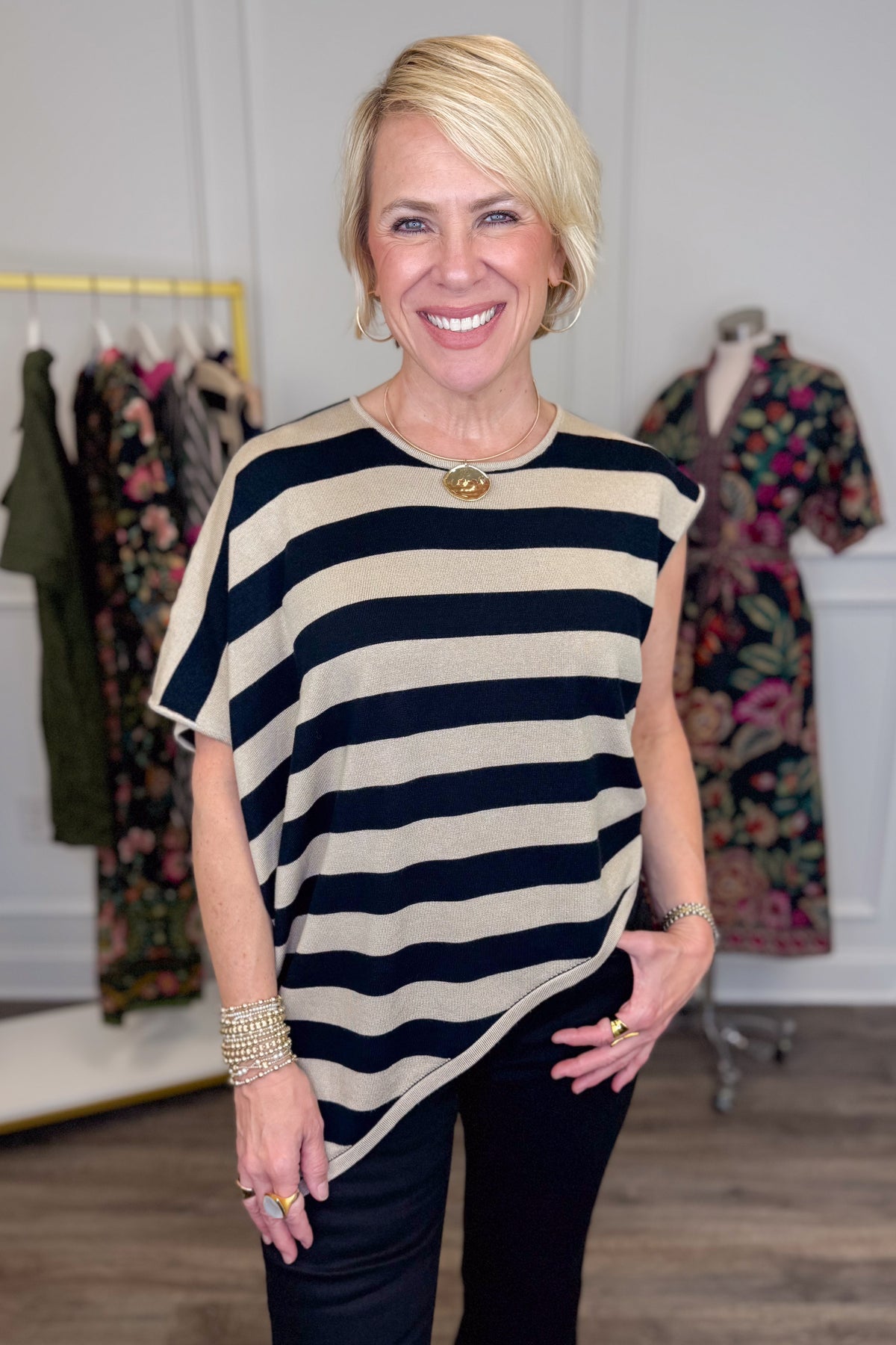 Front view of woman wearing Swagger Boutique’s Oatmeal &amp; Black Striped Asymmetrical Top with jeans with clothing hanging in the background.