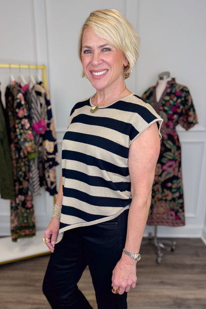Side view of woman wearing Swagger Boutique’s Oatmeal &amp; Black Striped Asymmetrical Top with jeans with clothing hanging in the background.