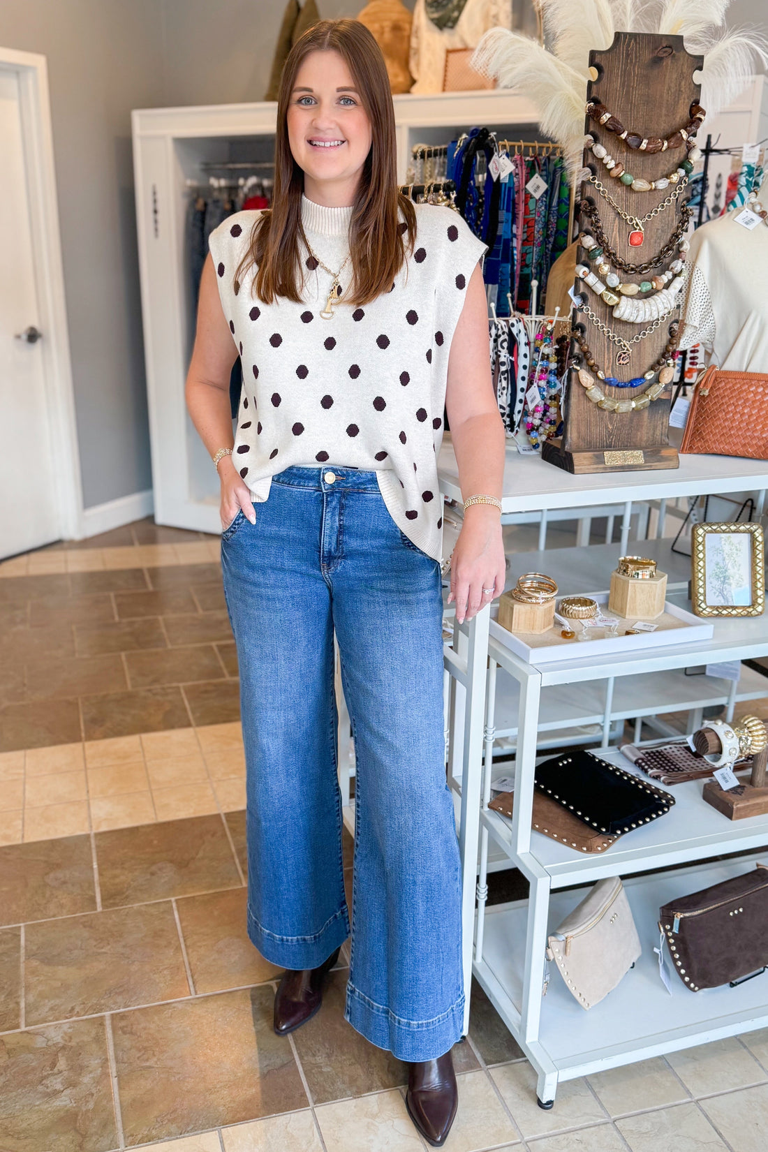 Front view of woman wearing Swagger Boutique’s Oatmeal &amp; Brown Polka Dot Sleeveless Sweater by Entro with jeans standing at the store.