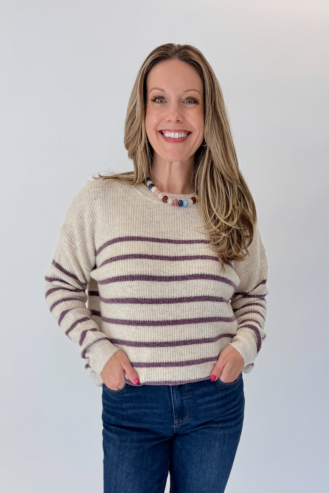 Front view of woman wearing Swagger Boutique’s Oatmeal &amp; Purple Striped Sweater with jeans against white studio background.