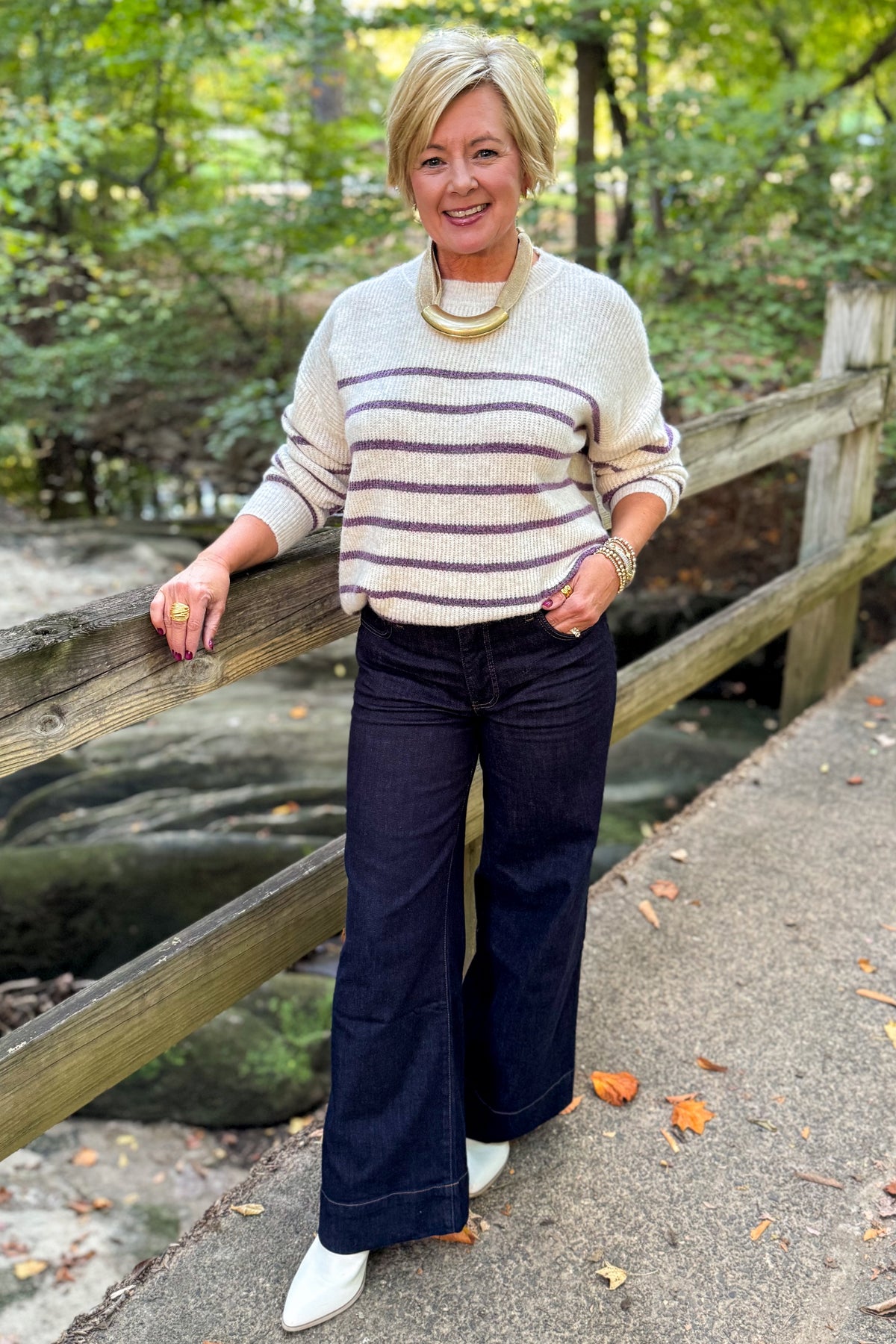 Front view of woman wearing Swagger Boutique’s Oatmeal &amp; Purple Striped Sweater with jeans standing outside on a bridge.
