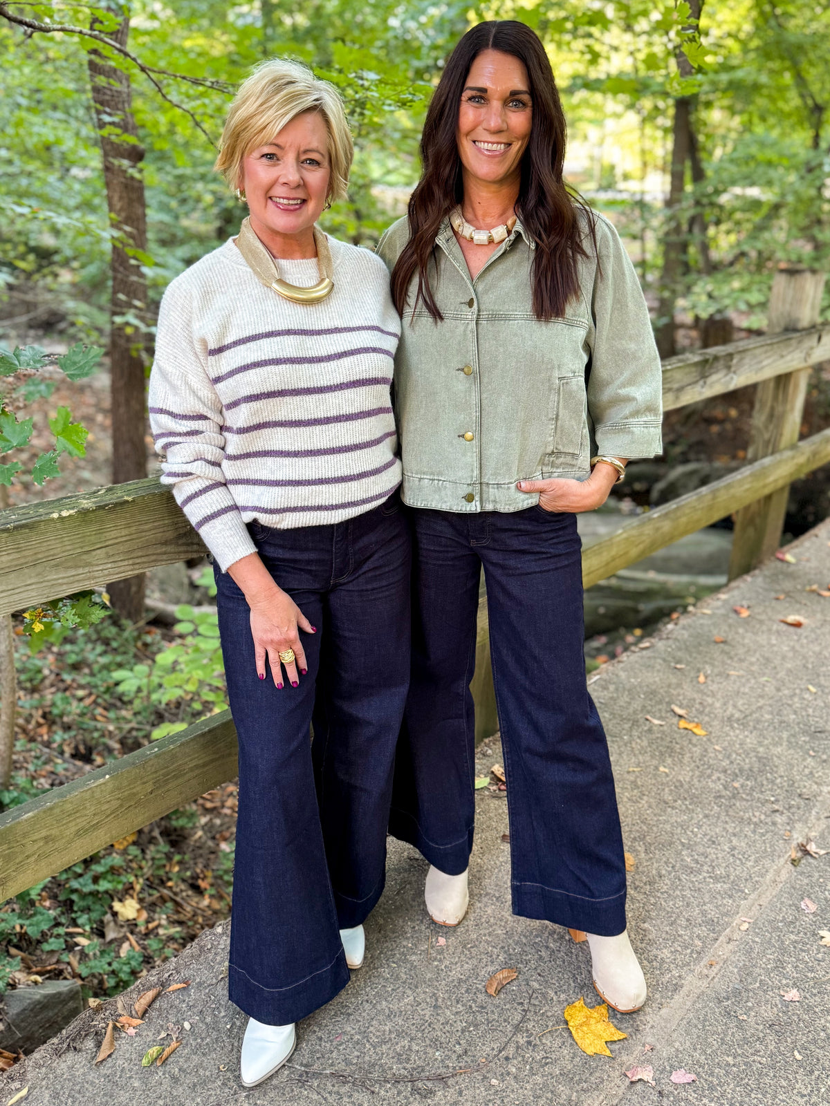 Front view of two women wearing Swagger Boutique’s Oatmeal &amp; Purple Striped Sweater &amp; Washed Olive Utility Jacket with jeans standing outside on a bridge. 