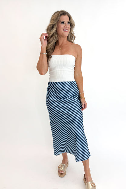 Front view of woman wearing Off White Ribbed Tube Top with Blue &amp; Navy Striped Satin Midi Skirt and sandals against white studio background.