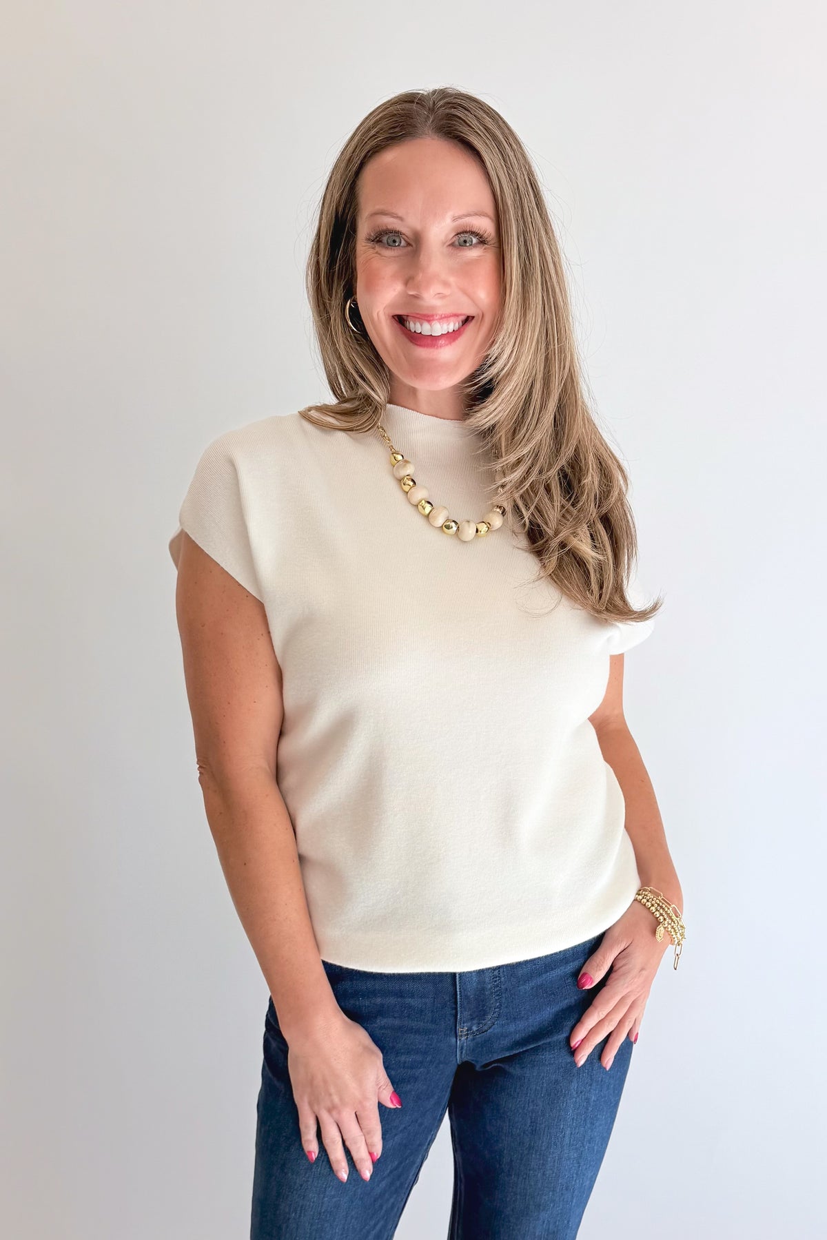 Front view of woman wearing Swagger Boutique’s Off White Shirred Cap Sleeve Sweater with jeans against white studio background.