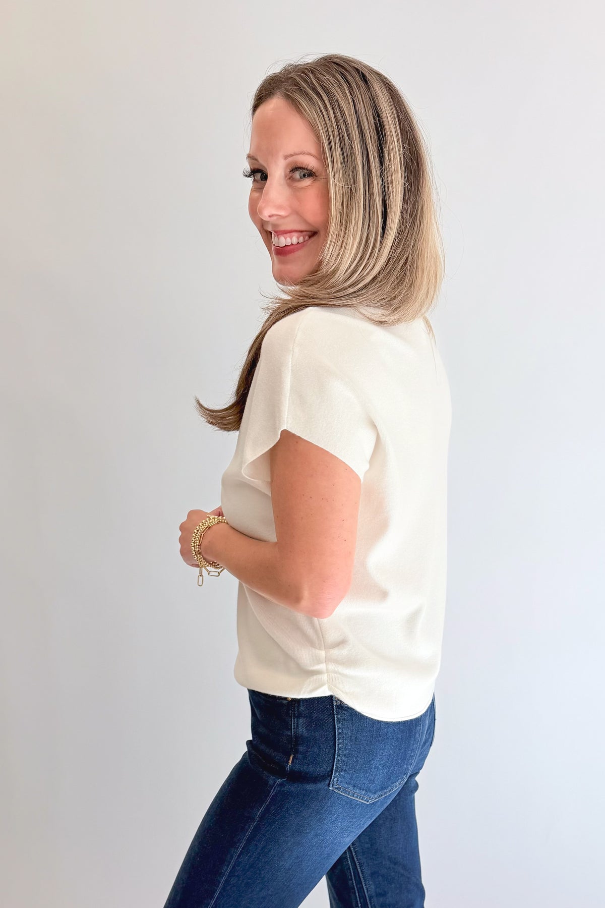 Side view of woman wearing Swagger Boutique’s Off White Shirred Cap Sleeve Sweater with jeans against white studio background.