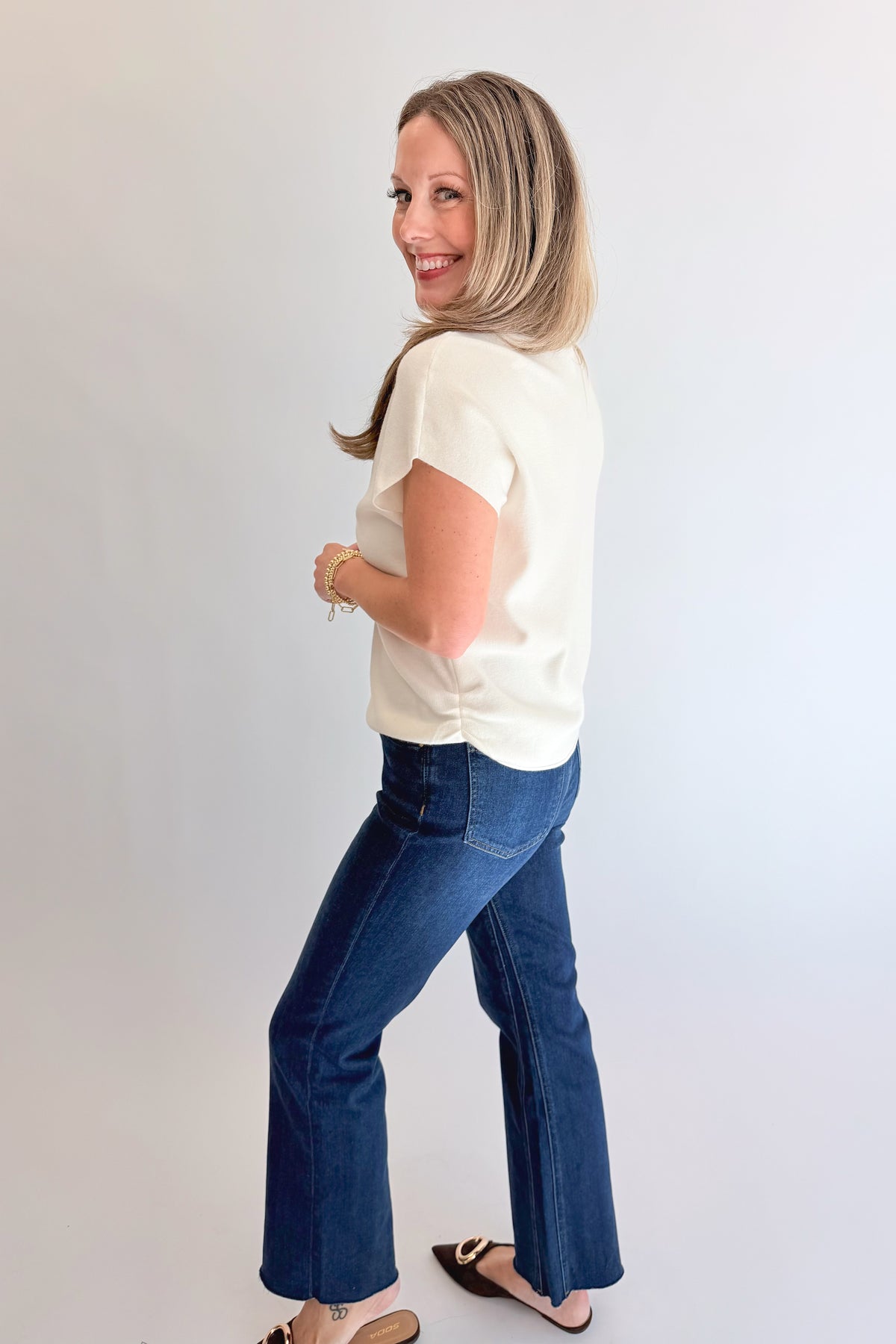 Side view of woman wearing Swagger Boutique’s Off White Shirred Cap Sleeve Sweater with jeans against white studio background.