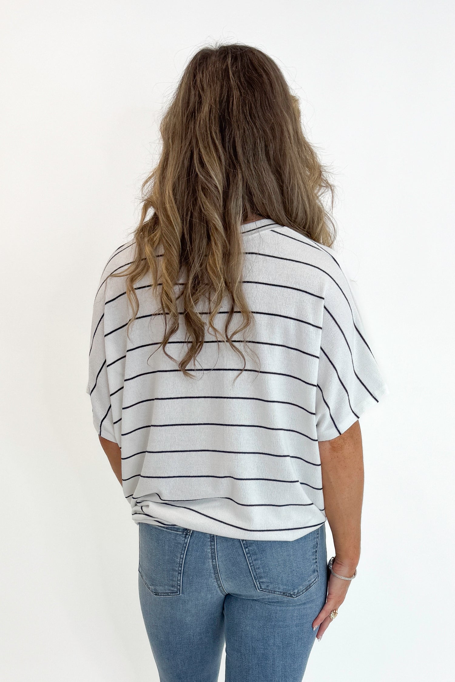 Back view of woman wearing Swagger Boutique’s Off White Striped Cocoon Top with jeans against white studio background.