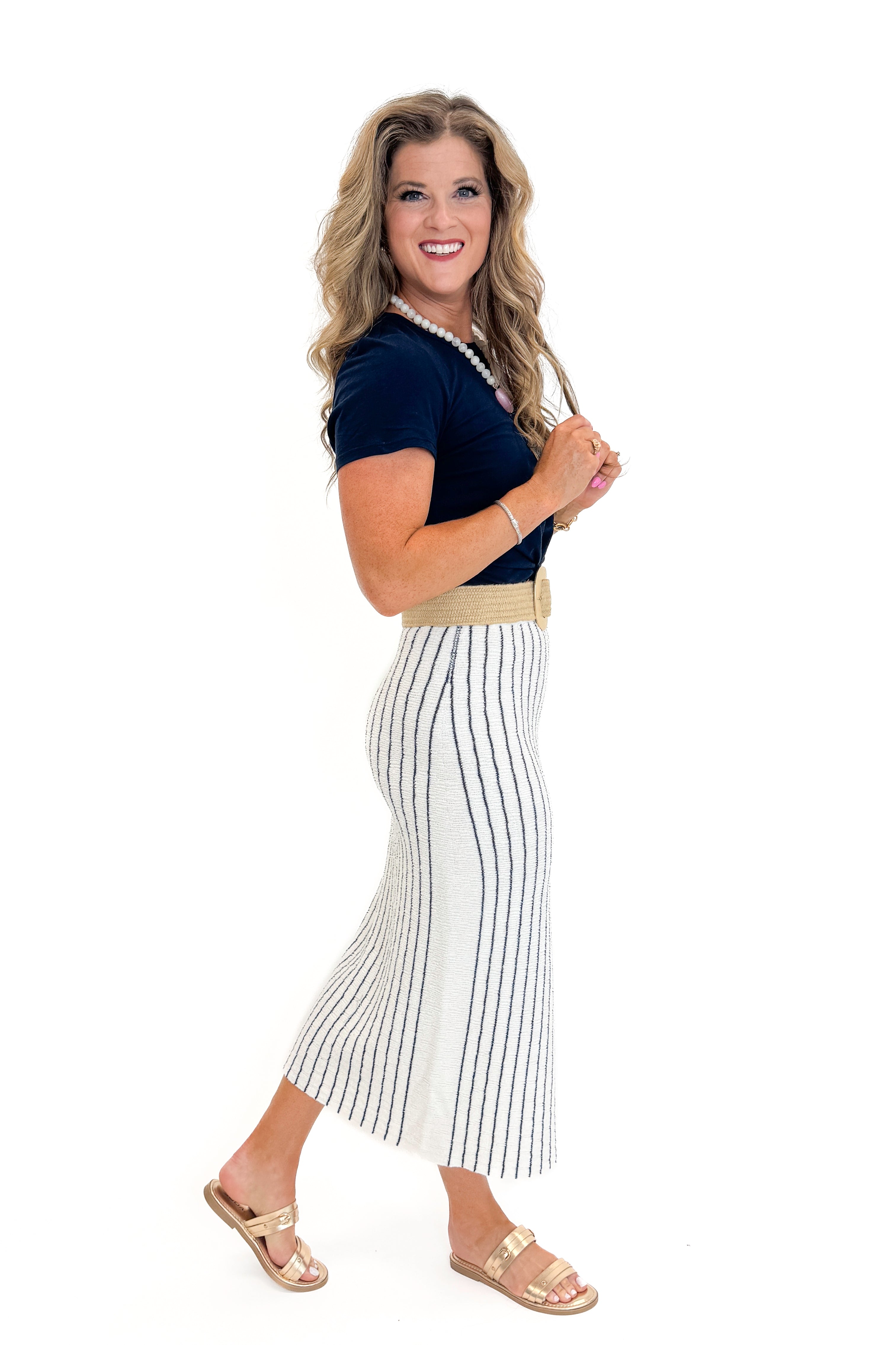 Side view of woman wearing Swagger Boutique’s Off White &amp; Navy Striped Midi Skirt With Slit and navy top against white studio background.