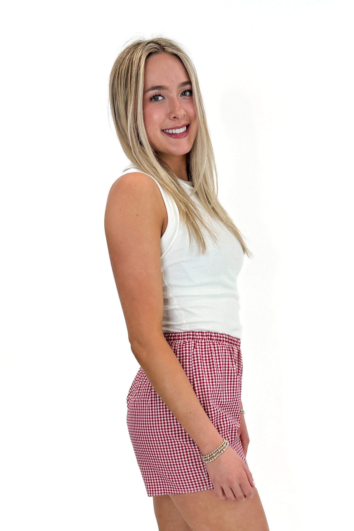 Side view of woman wearing Swagger Boutique’s Red Gingham Shorts with Off White Round Hem Ribbed Tank against white studio background.