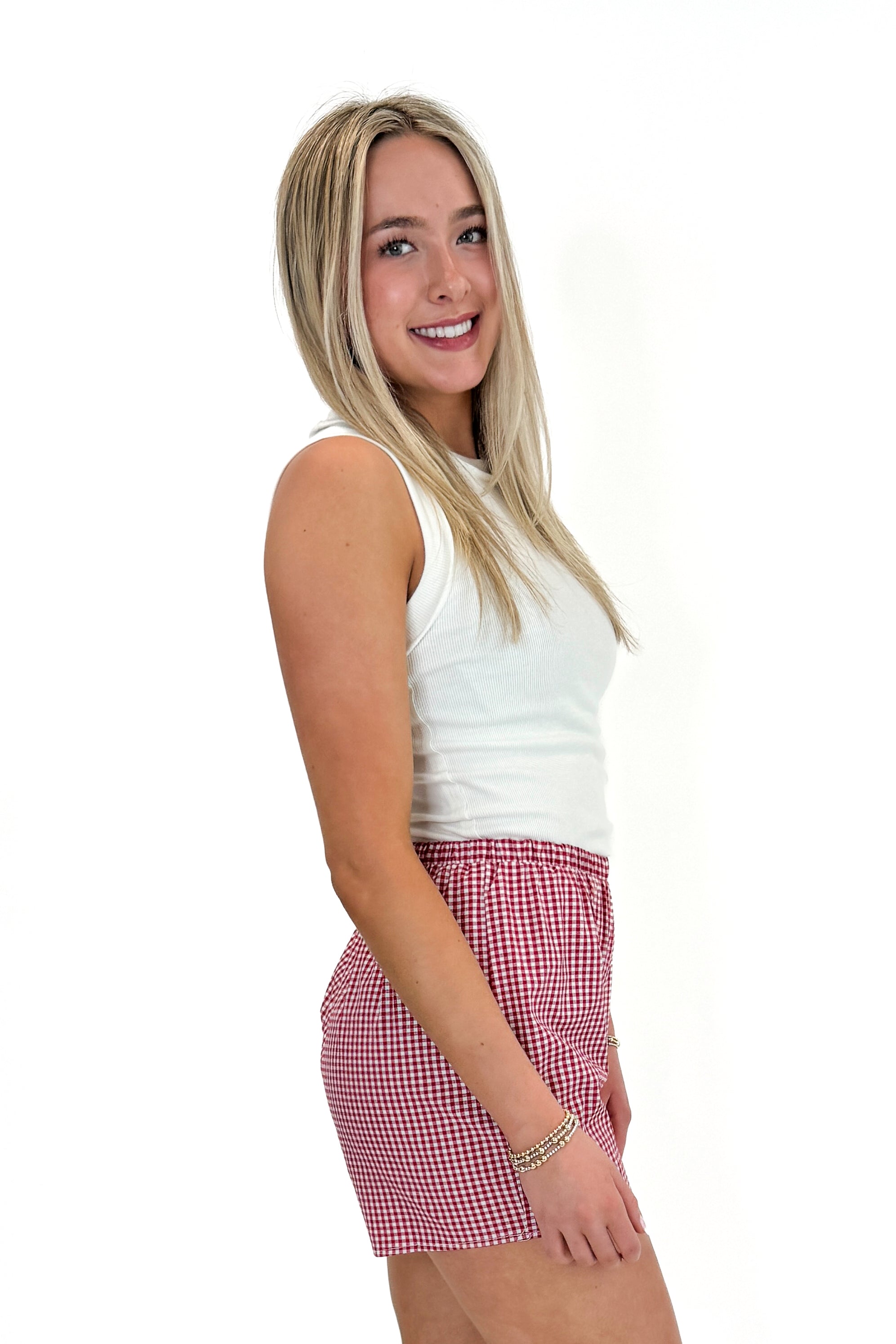 Side view of woman wearing Swagger Boutique’s Red Gingham Shorts with Off White Round Hem Ribbed Tank against white studio background.
