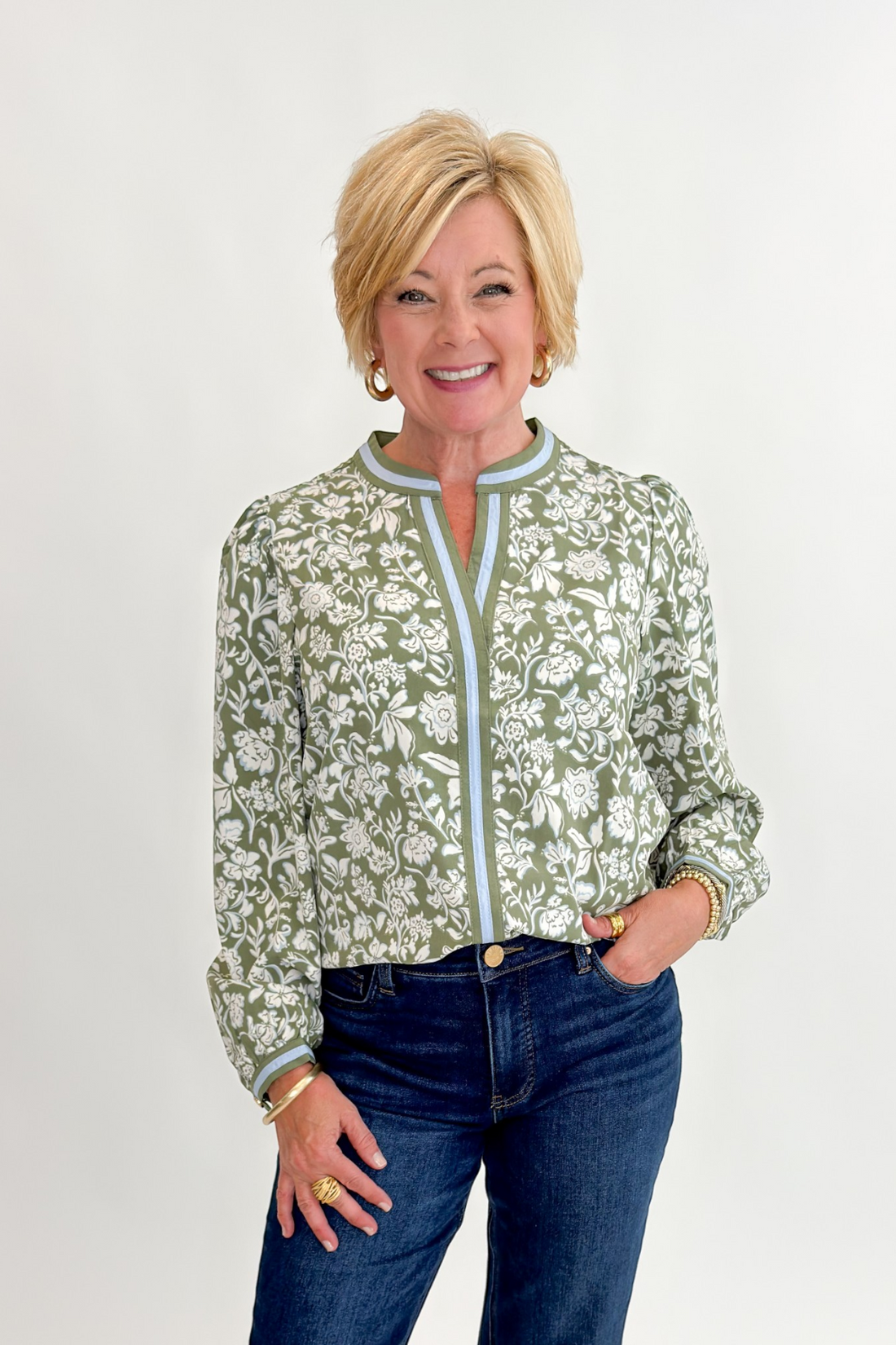 A smiling blonde woman wearing a sage green and white floral print long-sleeve blouse with light blue trim, paired with dark blue jeans, standing against a plain white studio background.