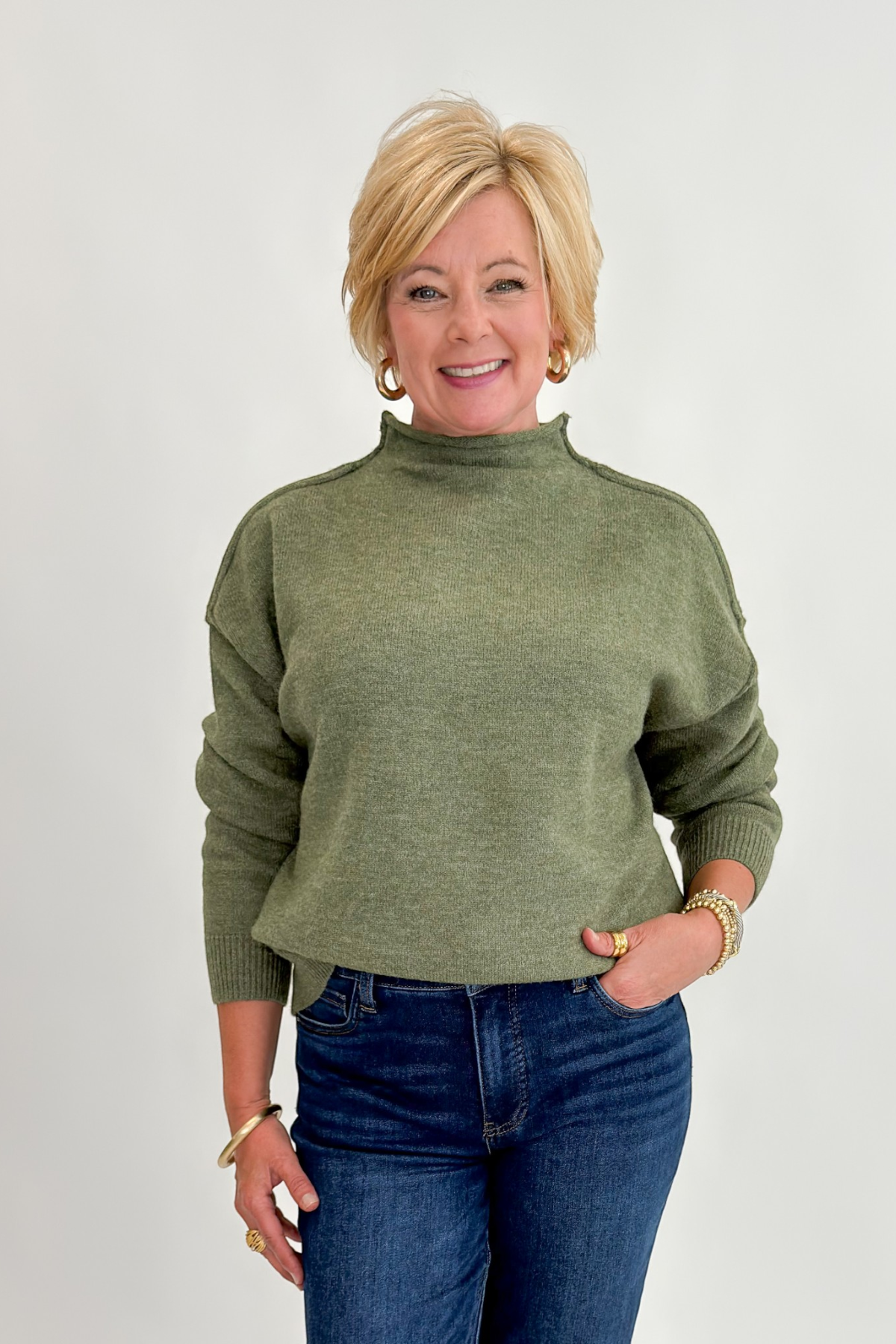 Front view of woman wearing Swagger Boutique’s Olive Mock Neck Sweater by Entro with jeans against white studio background.
