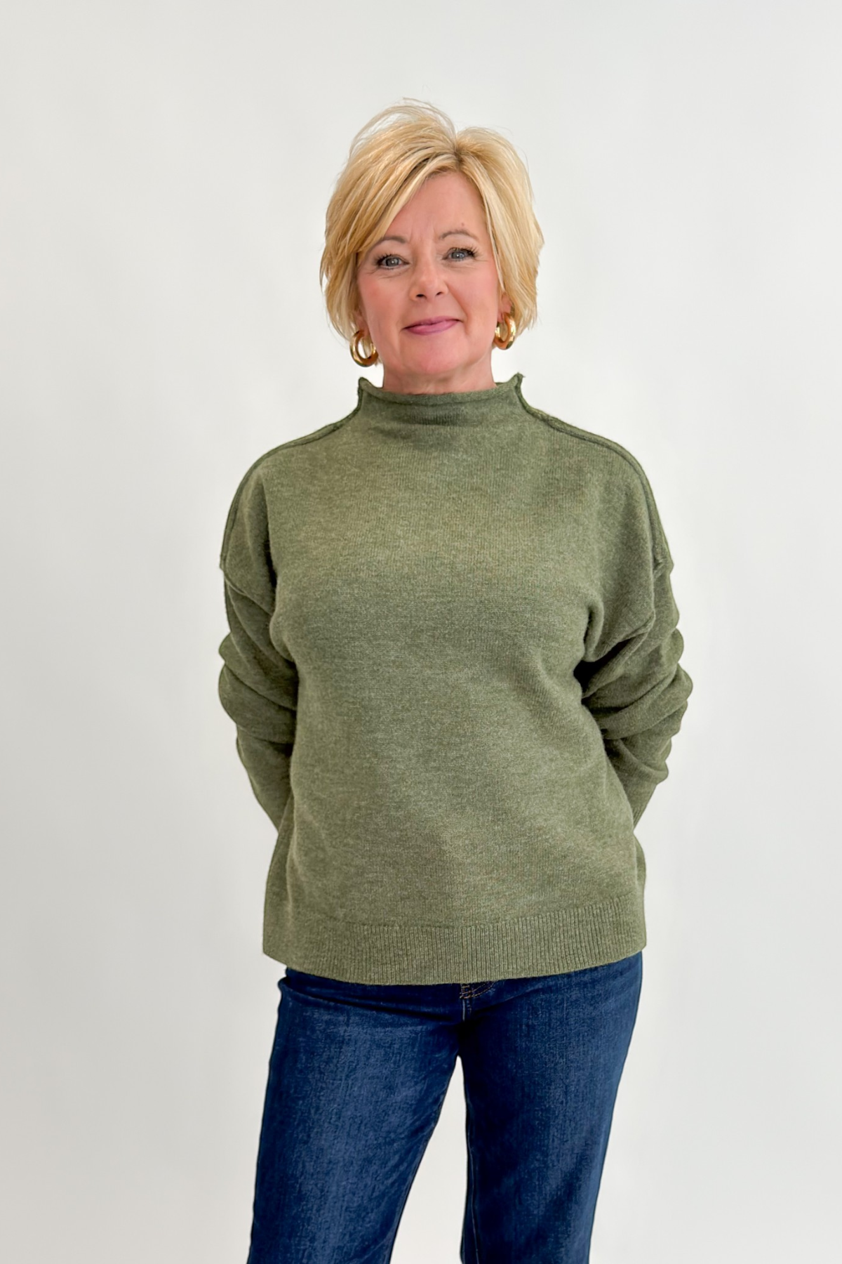 Front view of woman wearing Swagger Boutique’s Olive Mock Neck Sweater by Entro with jeans against white studio background.