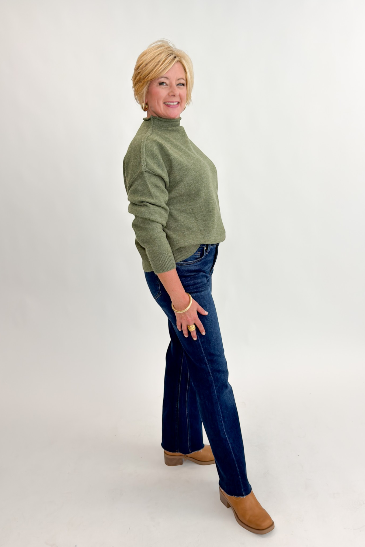 Side view of woman wearing Swagger Boutique’s Olive Mock Neck Sweater by Entro with jeans against white studio background.