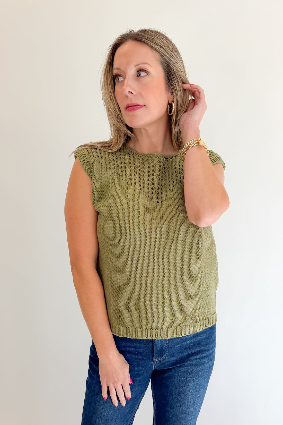 Front view of woman wearing Swagger Boutique’s Olive Open Knit Sleeveless Sweater with jeans against white studio background.