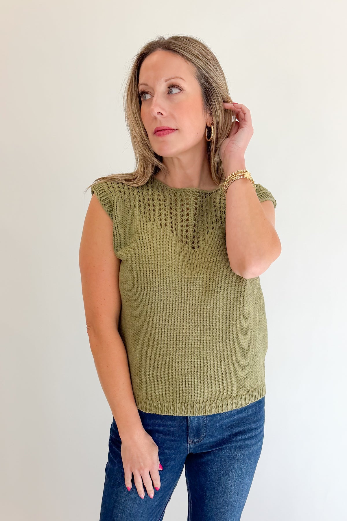 Front view of woman wearing Swagger Boutique’s Olive Open Knit Sleeveless Sweater with jeans against white studio background.