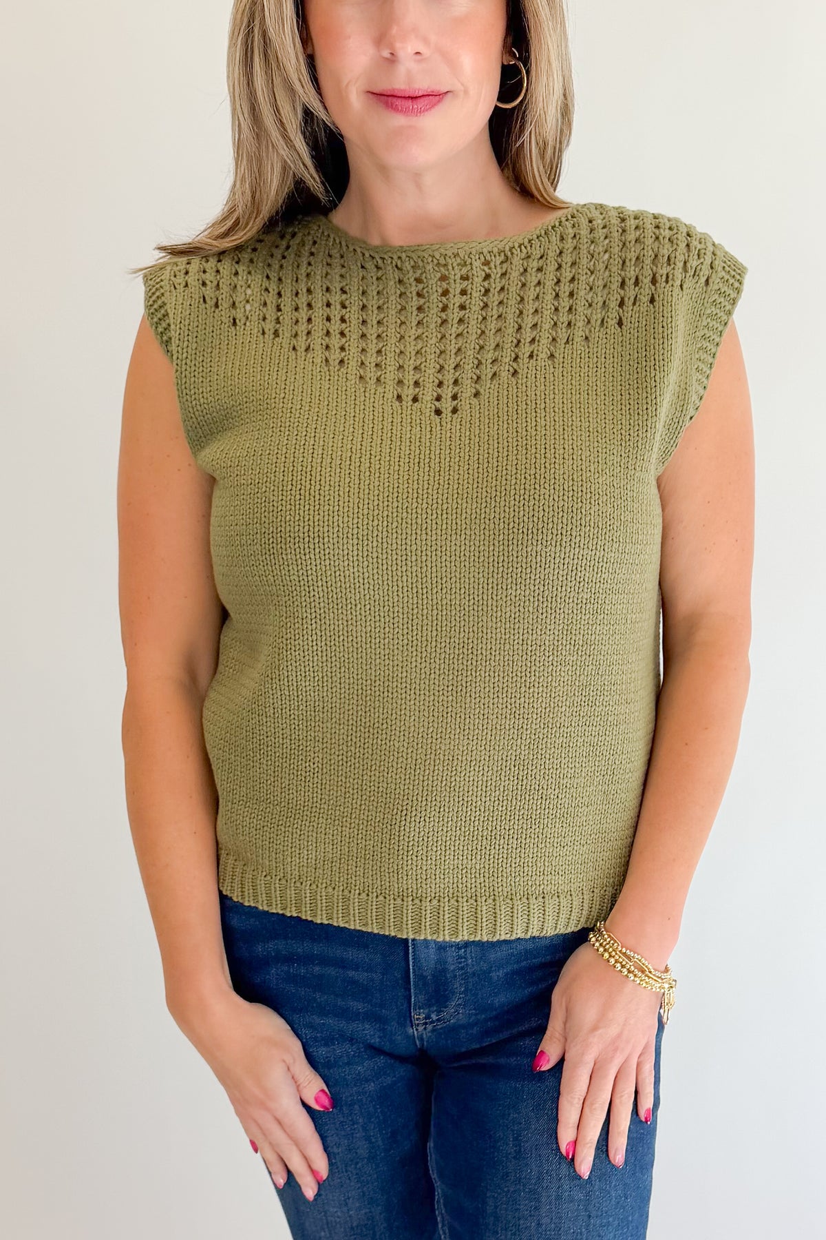 Front detail view of woman wearing Swagger Boutique’s Olive Open Knit Sleeveless Sweater with jeans against white studio background.