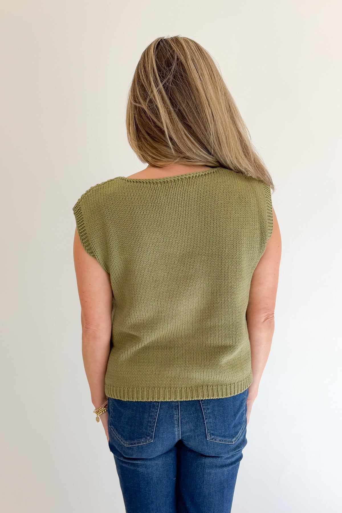 Back view of woman wearing Swagger Boutique’s Olive Open Knit Sleeveless Sweater with jeans against white studio background.