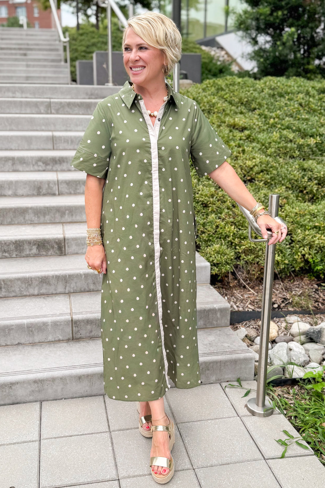 Front view of woman wearing Swagger Boutique’s Olive Polka Dot Midi Dress by Entro with sandals walking down a staircase outside.