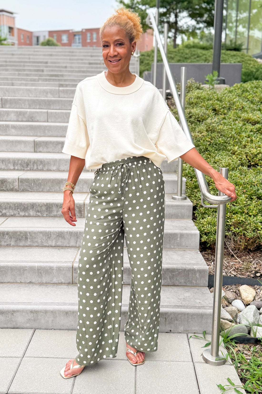 Front view of woman wearing Swagger Boutique’s Cream 3/4 Sleeve Lightweight Sweater by Entro with Olive Polka Dot Stripe Trim Pants by Entro standing by a staircase outside.