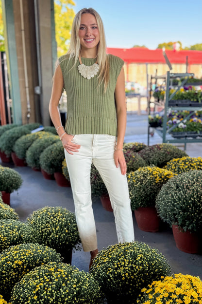 Front view of woman wearing Swagger Boutique’s Olive Ribbed Sleeveless Sweater with jeans at a garden center.