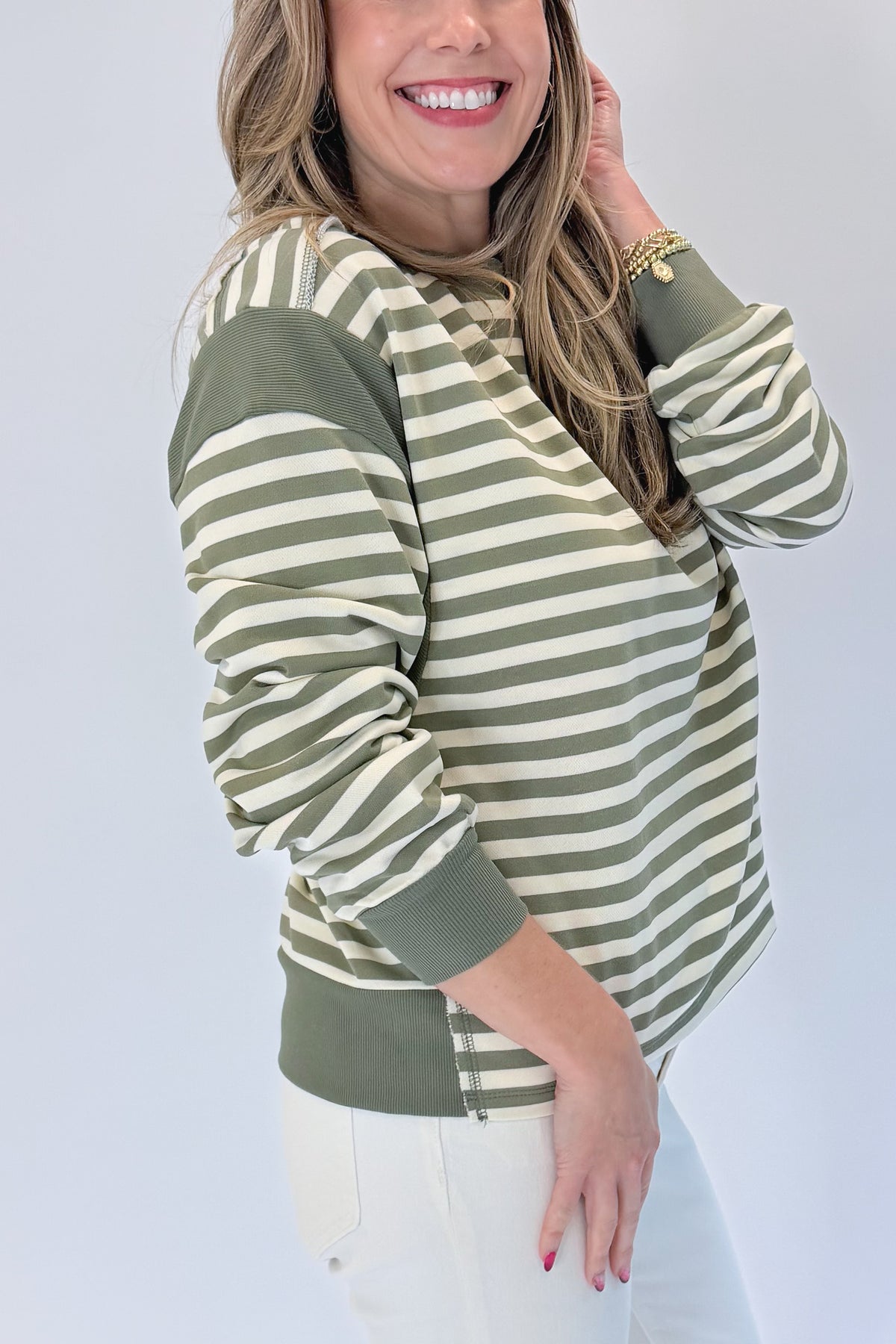 Side view of woman wearing Swagger Boutique’s Olive Striped Sweatshirt with jeans against white studio background.