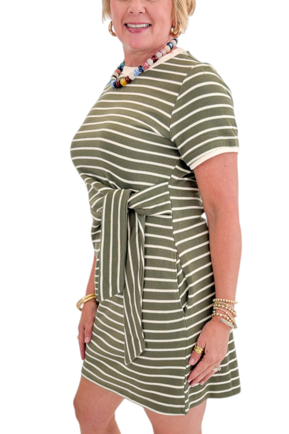 Side detail view of woman wearing Swagger Boutique’s Olive Striped Tie Waist Mini Dress by Entro with sneakers against white studio background.
