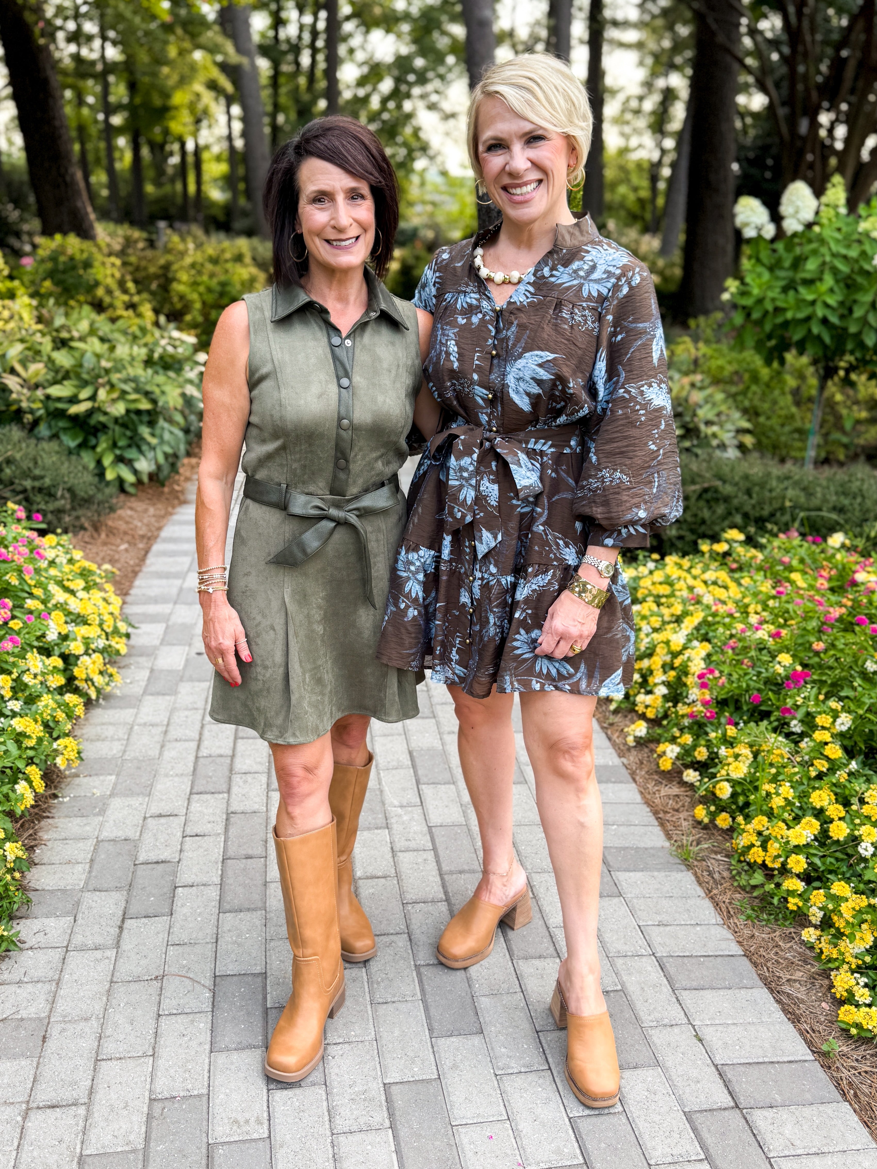 Front view of women wearing Swagger Boutique’s Brown &amp; Blue Floral Tie Waist Mini Dress and Olive Suede Dress standing outside by a garden.
