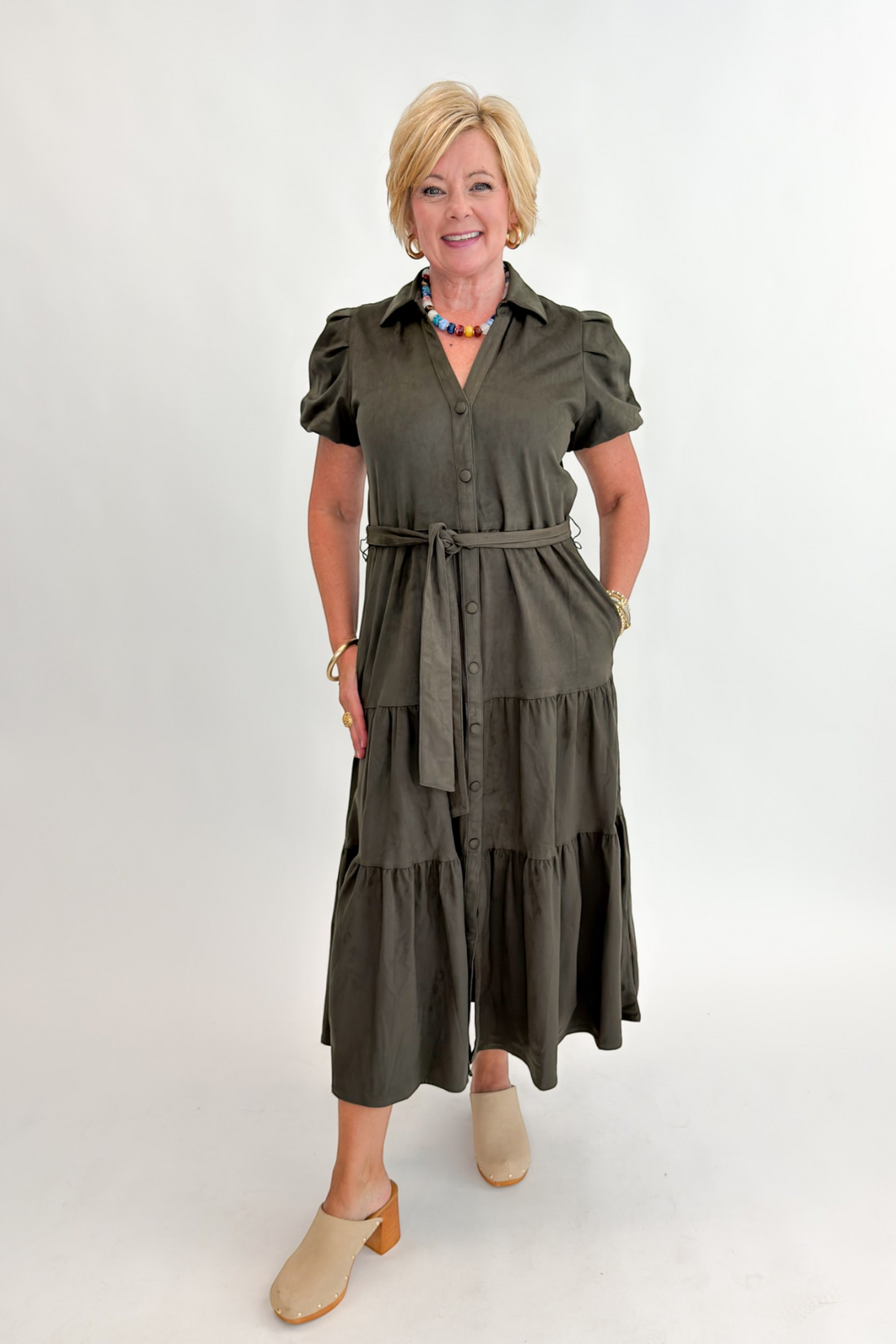Front view of woman wearing Swagger Boutique’s Olive Suede Tiered Midi Dress with clogs against white studio background.