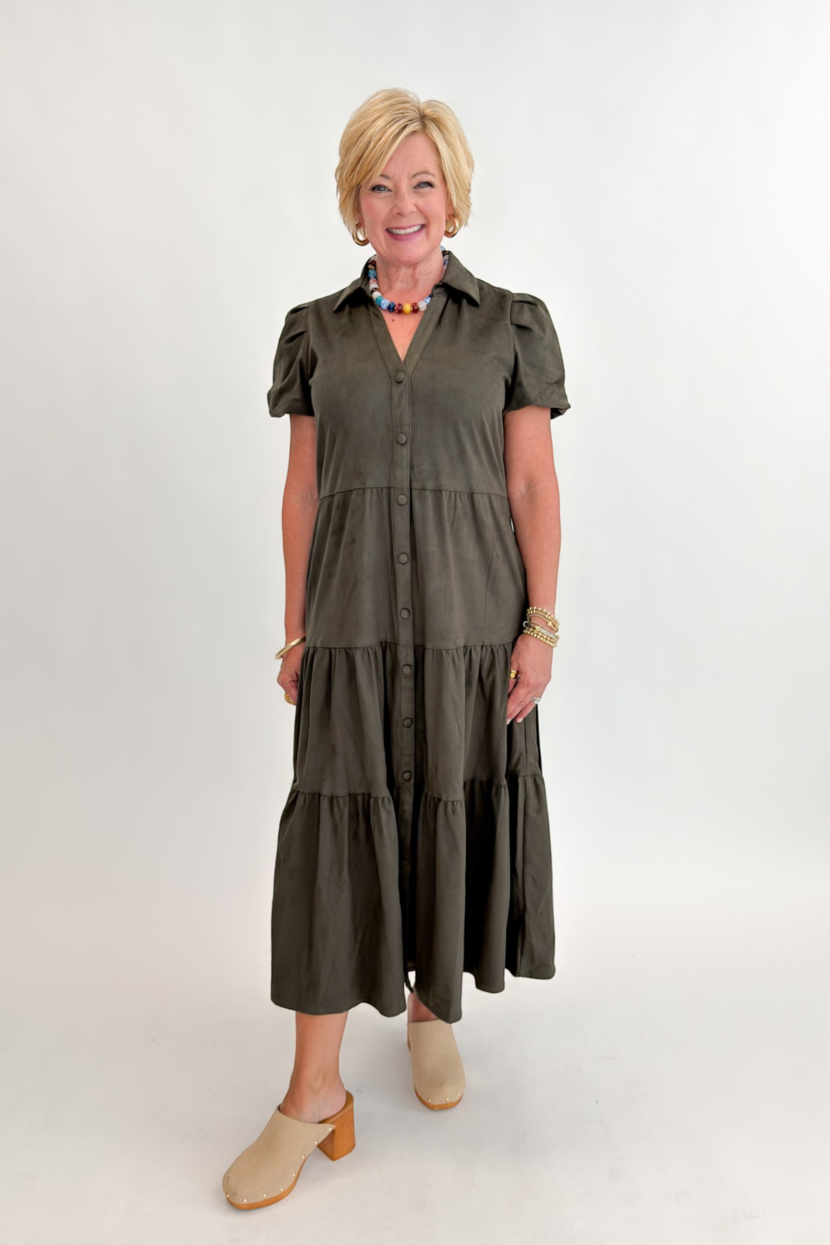 Front view of woman wearing Swagger Boutique’s Olive Suede Tiered Midi Dress with clogs against white studio background.