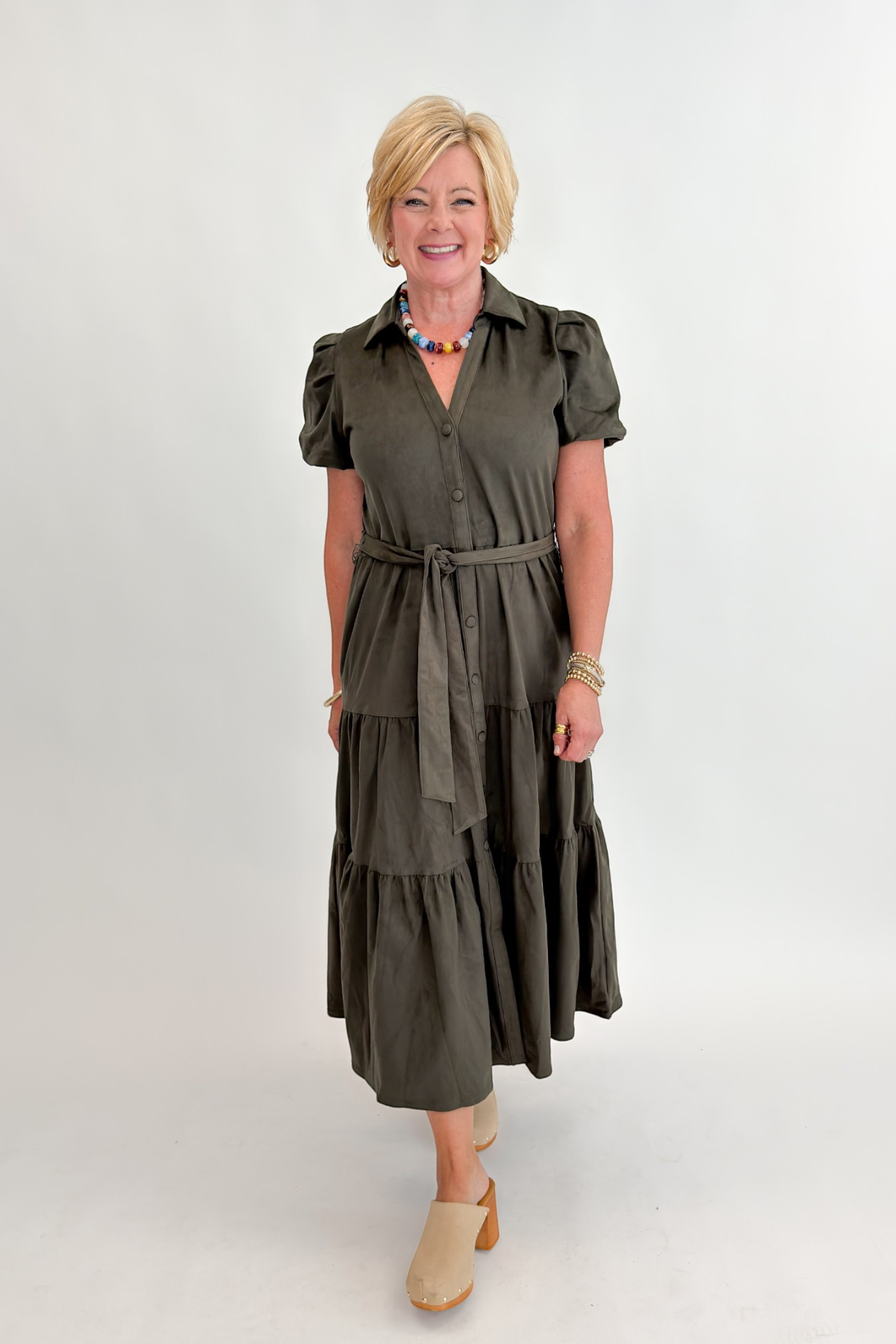 Front view of woman wearing Swagger Boutique’s Olive Suede Tiered Midi Dress with clogs against white studio background.