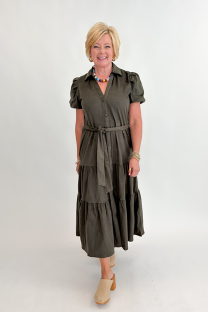 Front view of woman wearing Swagger Boutique’s Olive Suede Tiered Midi Dress with clogs against white studio background.