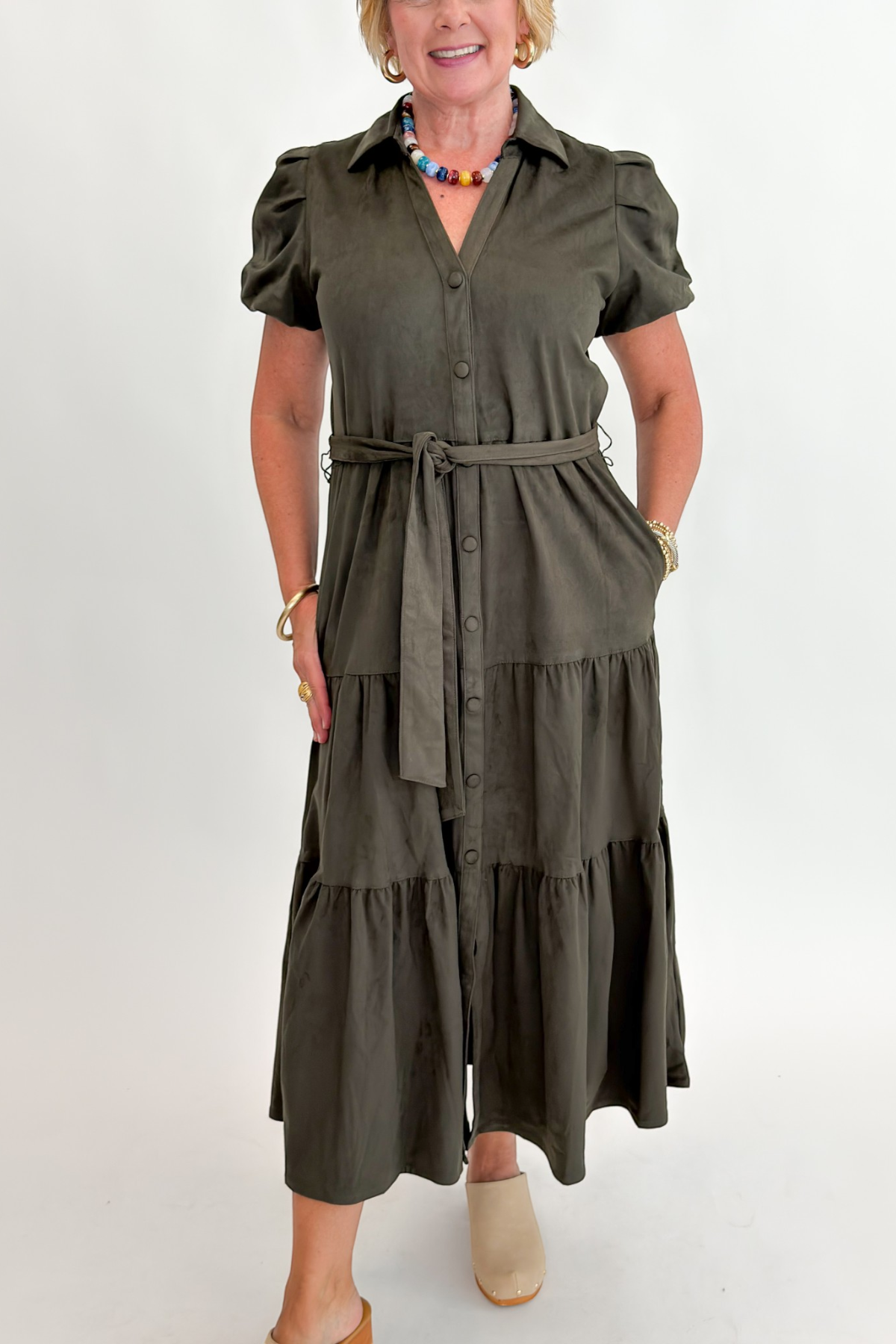 Front detail view of woman wearing Swagger Boutique’s Olive Suede Tiered Midi Dress with clogs against white studio background.