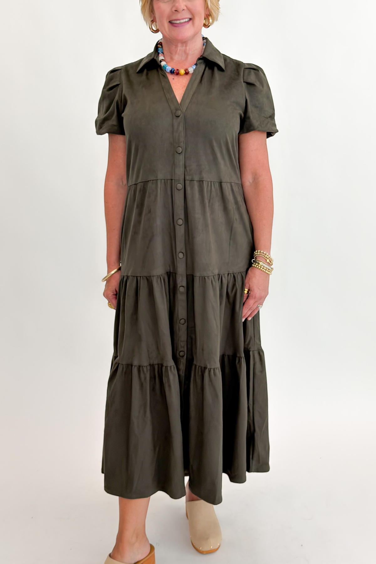 Front detail view of woman wearing Swagger Boutique’s Olive Suede Tiered Midi Dress with clogs against white studio background.