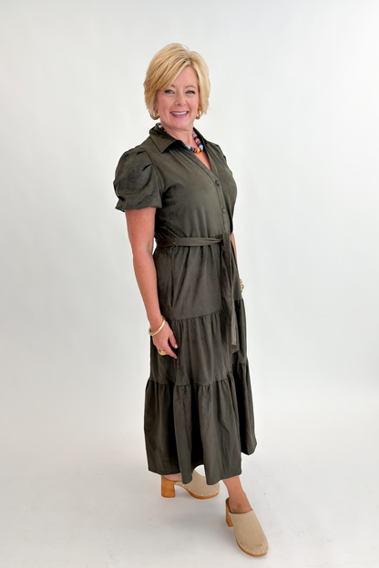 Side view of woman wearing Swagger Boutique’s Olive Suede Tiered Midi Dress with clogs against white studio background.