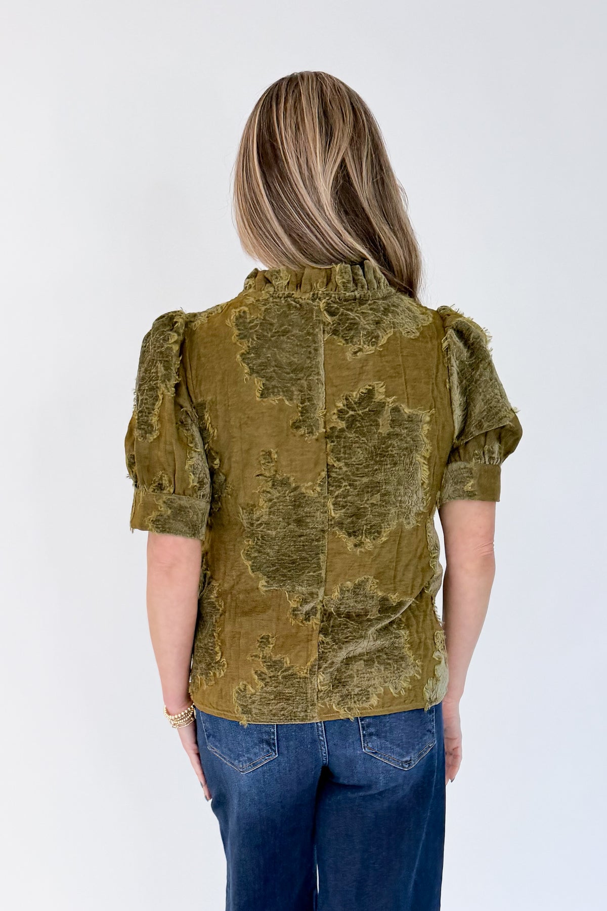 Back view of woman wearing Swagger Boutique’s Olive Velvet Ruffle Neck Top with jeans against white studio background.